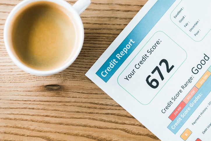 A white cup full of coffee on a wood table next to a piece of paper with “credit report” and “your credit score: 672” written on it.