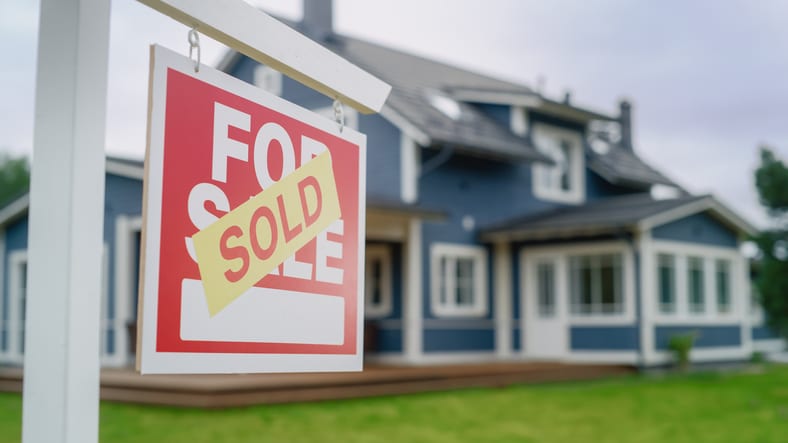 Close-up of a “For Sale” sign with a “Sold” sticker on it on the lawn of a blue house with white doors and borders around the windows.