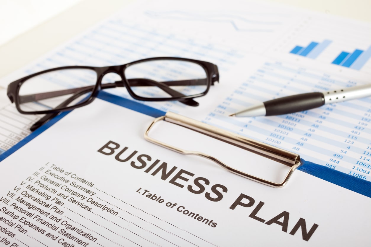 Glasses and a pen next to a blue clipboard with a business plan on it over a spreadsheet on a desk.